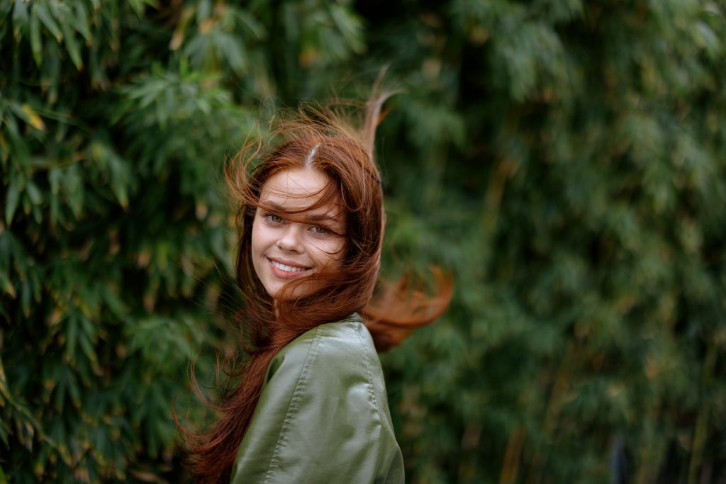 Portrait of a beautiful young girl in the city with a beautiful smile and red hair in a green raincoat in the city against a background of bamboo in spring, lifestyle in the city. High quality photo